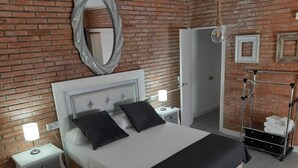 Une chambre avec un mur en briques, un lit avec une tête de lit blanche, deux oreillers noirs, une table de chevet avec une lampe et une armoire.