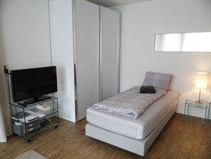 Une chambre à coucher de taille modeste, avec un lit simple, un téléviseur posé sur un meuble en verre, une lampe de chevet et une armoire.