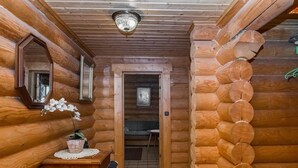 Interior de una cabaña de troncos con techo de madera, un candelabro, un espejo, una mesita con una planta y una puerta que da a otra habitación.