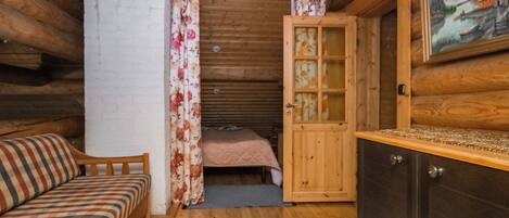 A cozy cabin interior with a wooden ceiling, a bed with floral curtains, a small kitchenette, and a painting on the wall.