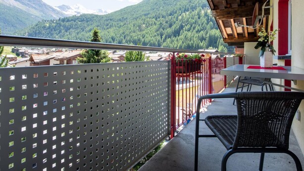 A balcony with a view of a mountain landscape, a table with chairs, and a red and white wall.