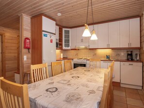 Een kleine keuken met houten kastjes, een eettafel met een tafelkleed met klokken, en een inbouwvaatwasser.
