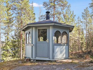 Una cabaña pequeña de un piso con chimenea, ventanas grandes y una puerta, rodeada por un bosque.
