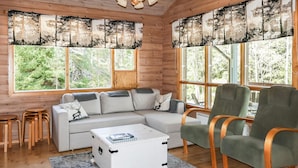 A log cabin interior with a sectional sofa, a green armchair, a white chest of drawers, and large windows with forest views. - Leijankorpi by Interhome (Pöytyä)