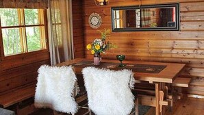 A wooden cabin interior with a dining table, two white furry chairs, a vase with flowers, a clock, and a deer head mounted on the wall.