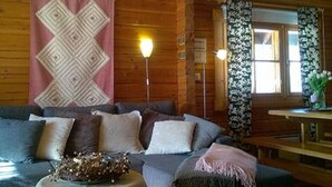 A cozy cabin interior with a wooden ceiling, a patterned wall hanging, a sofa with cushions, a small table with a lamp, and a window with curtains.