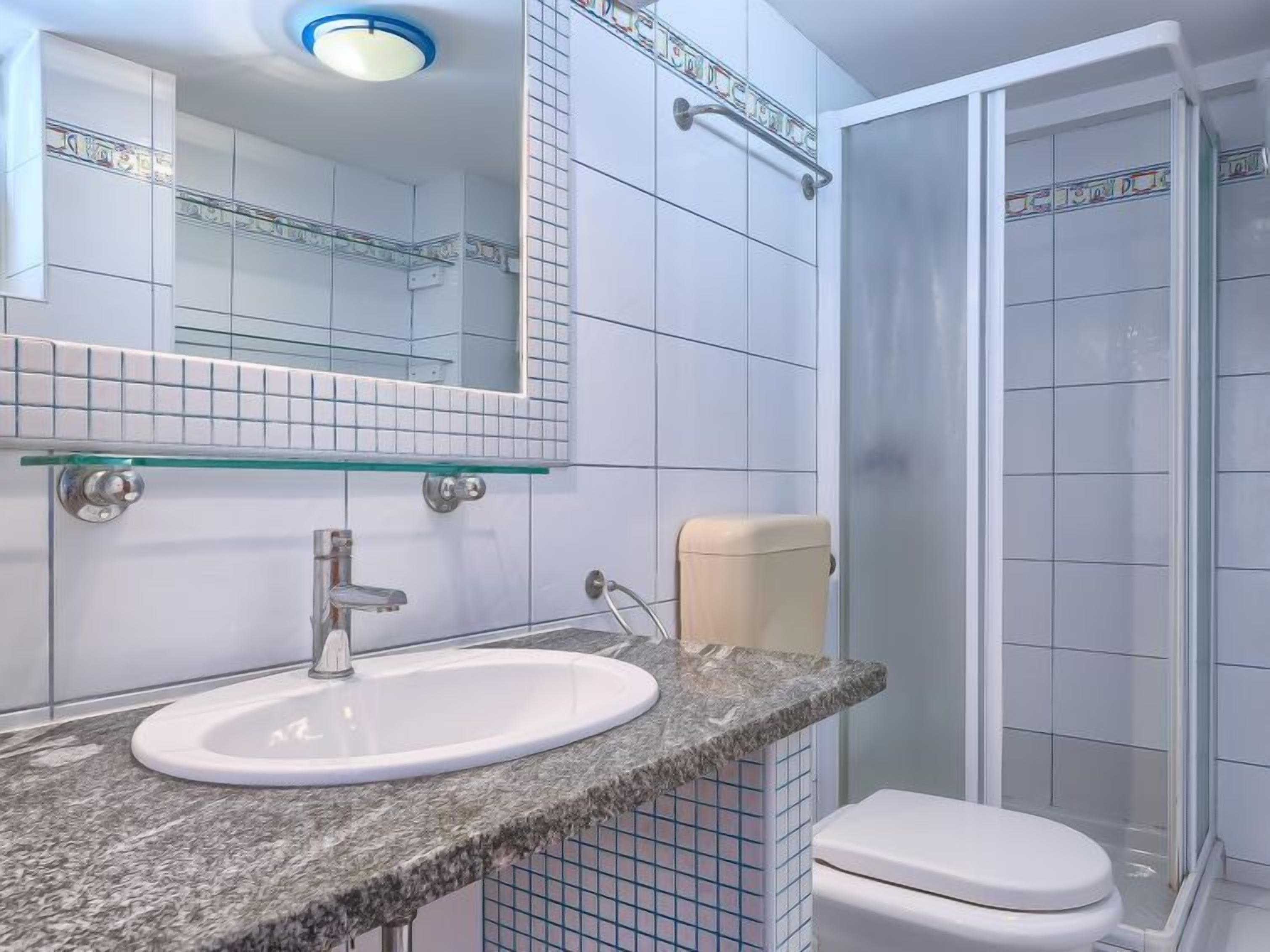 A bathroom with a white sink, a glass shelf, a toilet, and a shower area.