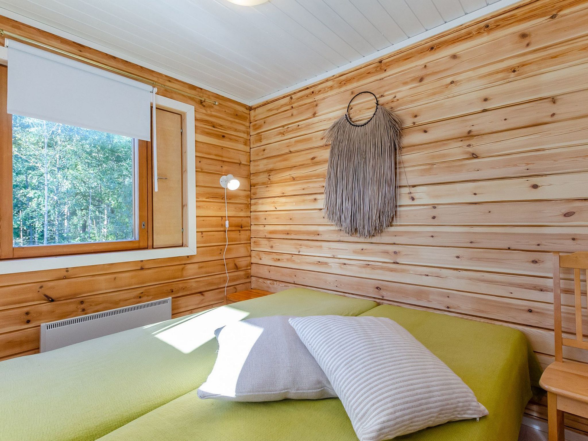 A room with wooden walls, a bed with green bedding, a window with a view of trees, and a wall-mounted light fixture.