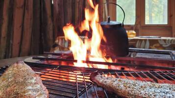 A grill with two pieces of meat cooking over an open flame, with a pot on a separate burner in the background.