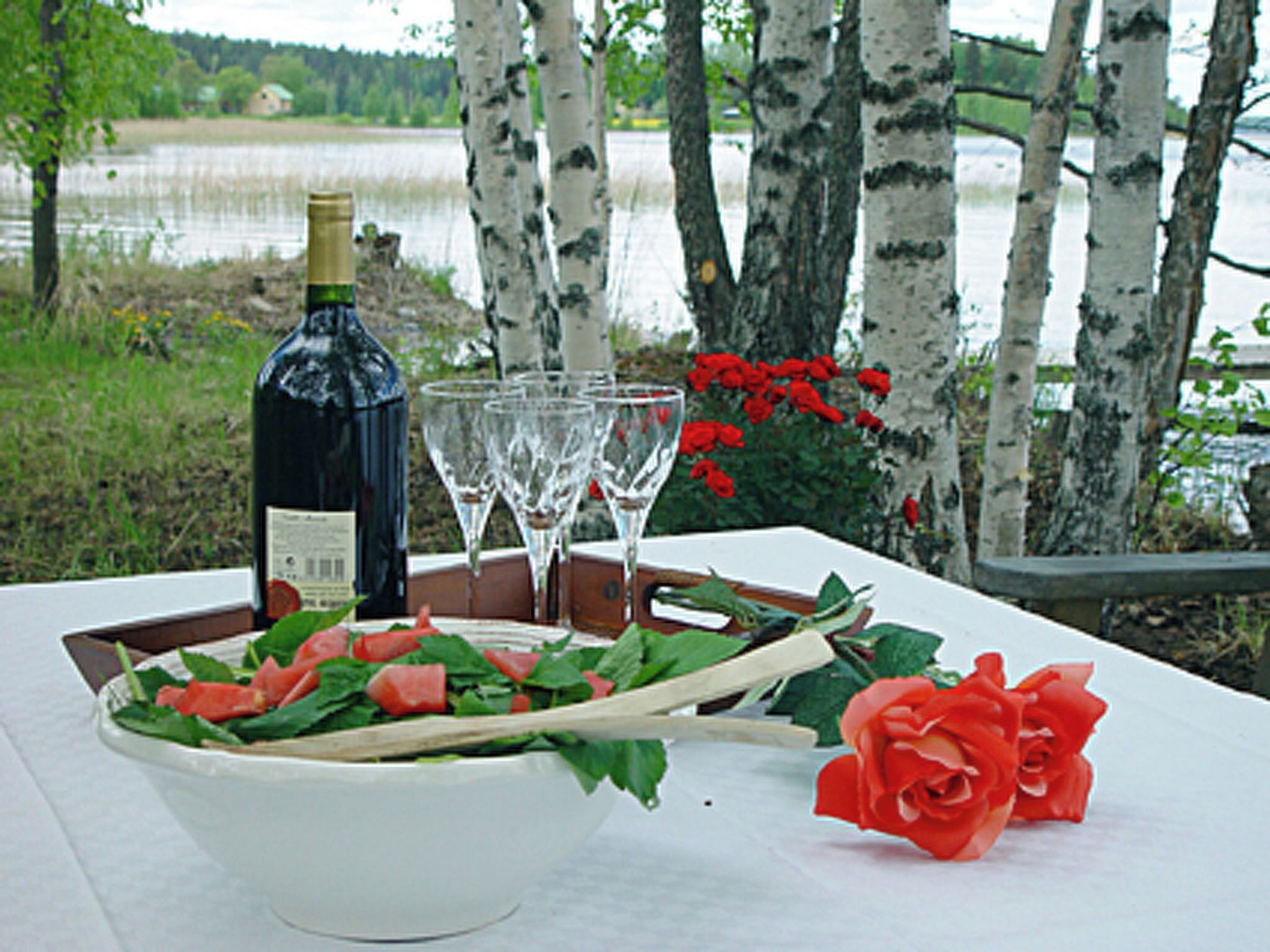 A table with a bottle of wine, glasses, a bowl of salad, and a rose.