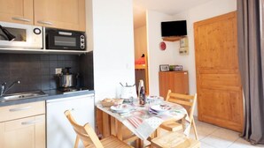 A compact kitchen with wooden cabinets, a microwave, and a stove. There is a dining table with chairs and a TV mounted on the wall. - Plein Soleil by Interhome (La Toussuire)