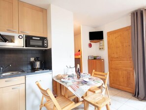 A compact kitchen with wooden cabinets, a microwave, and a stove. There is a dining table with chairs and a TV mounted on the wall.