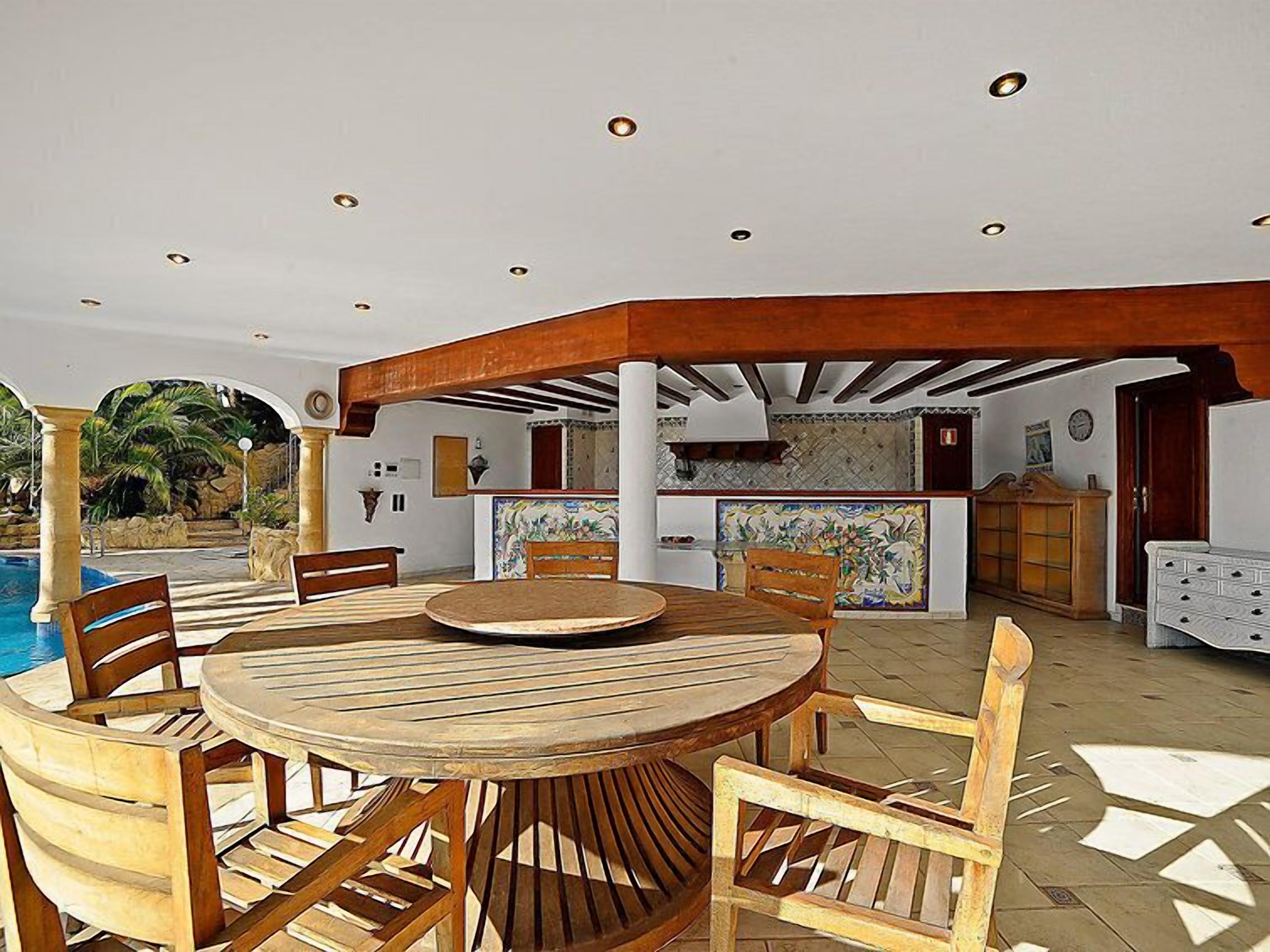 An outdoor dining area with a round wooden table and chairs, a pool, and a kitchen in the background.