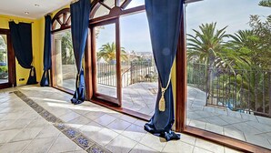 A spacious balcony with large windows and curtains, offering a view of palm trees and a distant cityscape. - St Anthony by Interhome (Jávea)