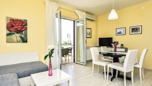 A modern living room with a grey sofa, a white table, and a dining area with white chairs. There is a painting of a rose on the wall and a vase with flowers on the table. - Apartment Marija by Interhome (Pag/Dinjiška)