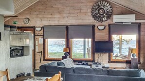L’intérieur chaleureux d’un chalet avec un plafond en bois, un canapé, une table à manger avec des chaises, une cheminée et de grandes fenêtres donnant sur un plan d’eau.