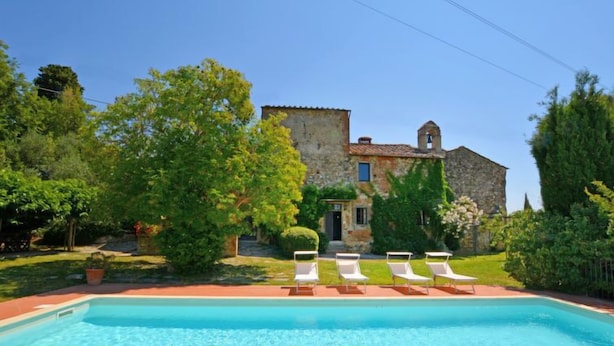 Una piscina con lettini, immersa nel verde e affiancata da un edificio in pietra con un campanile.