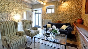 Un accogliente soggiorno con pareti in pietra, soffitto in legno, una poltrona a righe, un divano nero e un tavolino in vetro con oggetti decorativi.