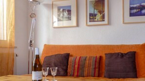 A room with an orange sofa, two framed pictures on the wall, a bottle of wine, and two glasses on a wooden table. - Hauts de Morthey No 33 by Interhome (Ovronnaz)