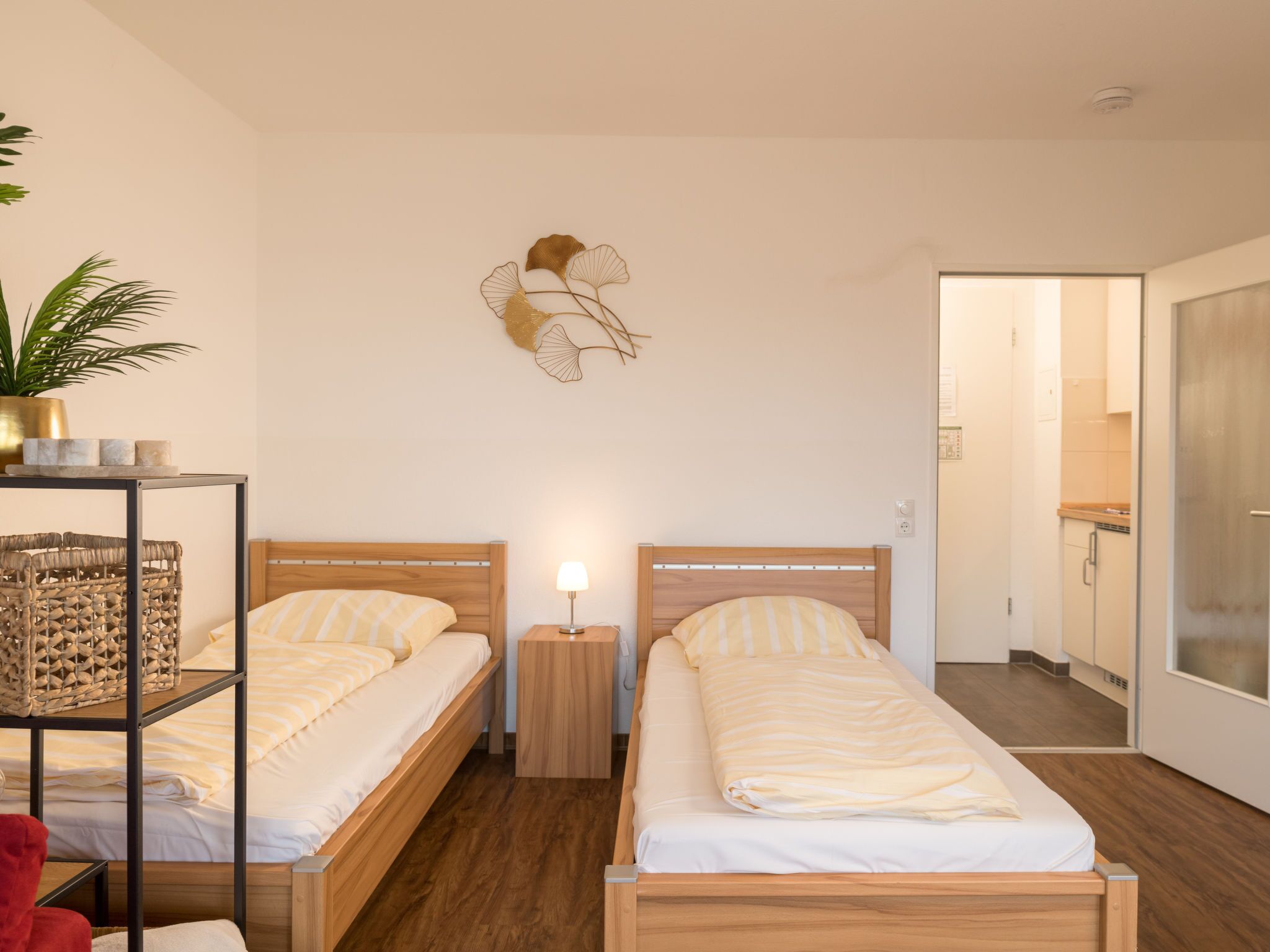 A modern hotel room with two wooden beds, a small bedside table with a lamp, a wall-mounted decorative piece, and a view into the adjacent kitchen.