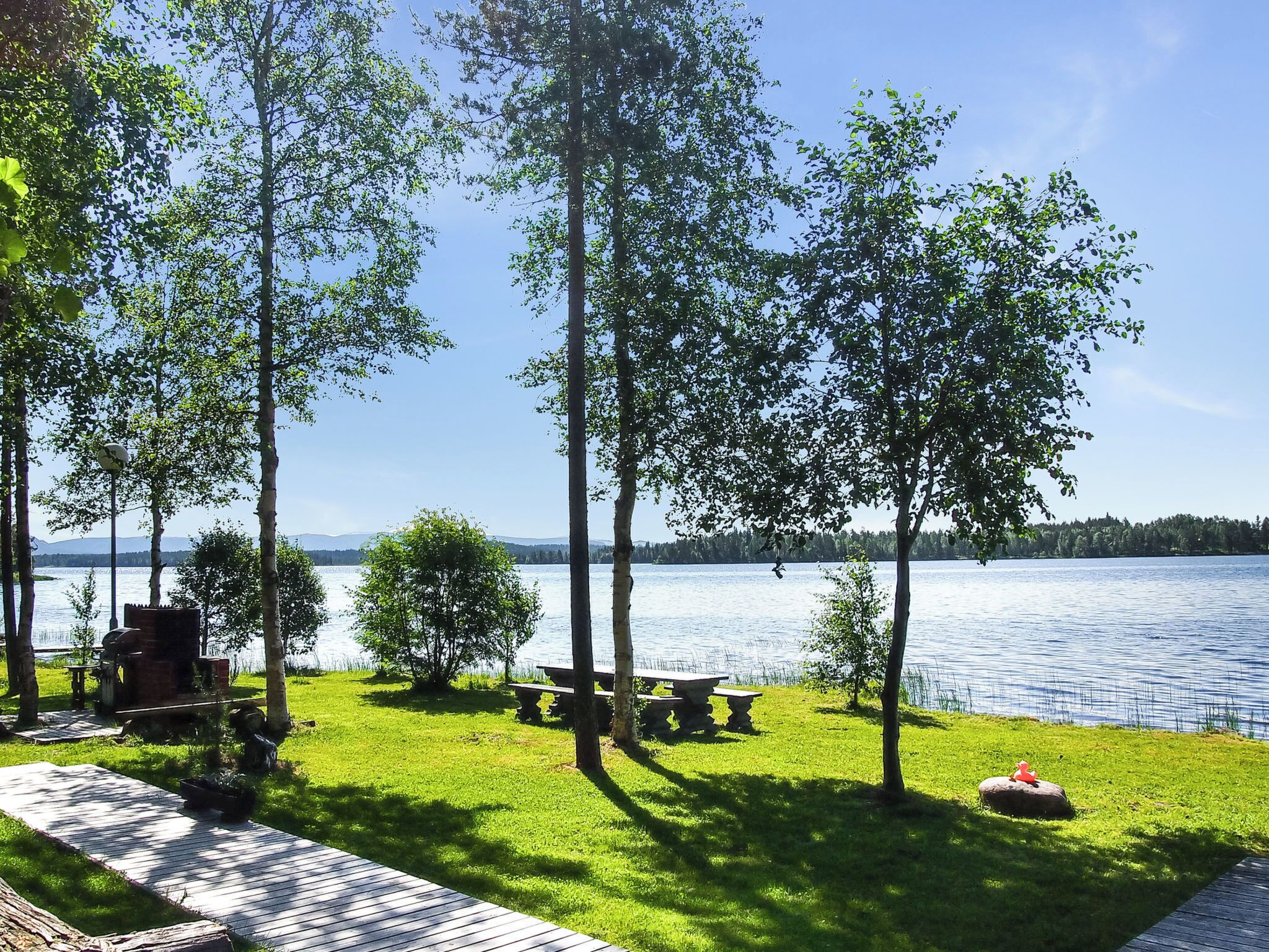 A lakeside patio with picnic tables and a grill, surrounded by trees.