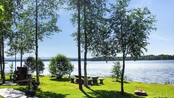 A lakeside patio with picnic tables and a grill, surrounded by trees.