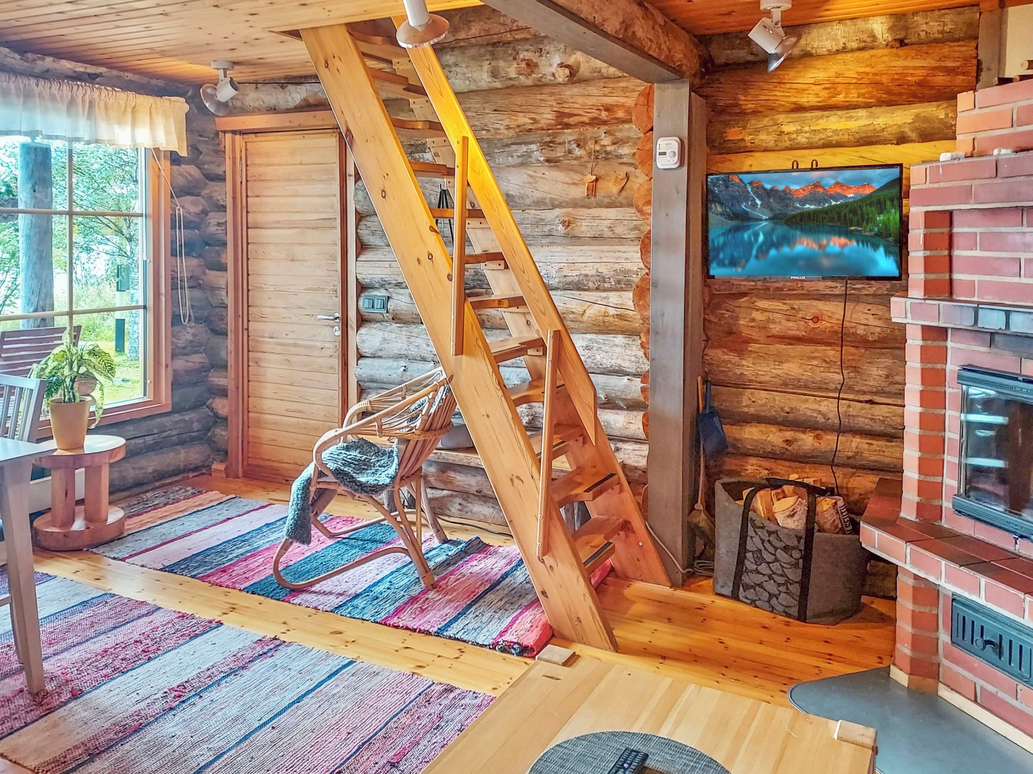 A log cabin interior with a wooden staircase, a rocking chair, a fireplace, and a television mounted on the wall.
