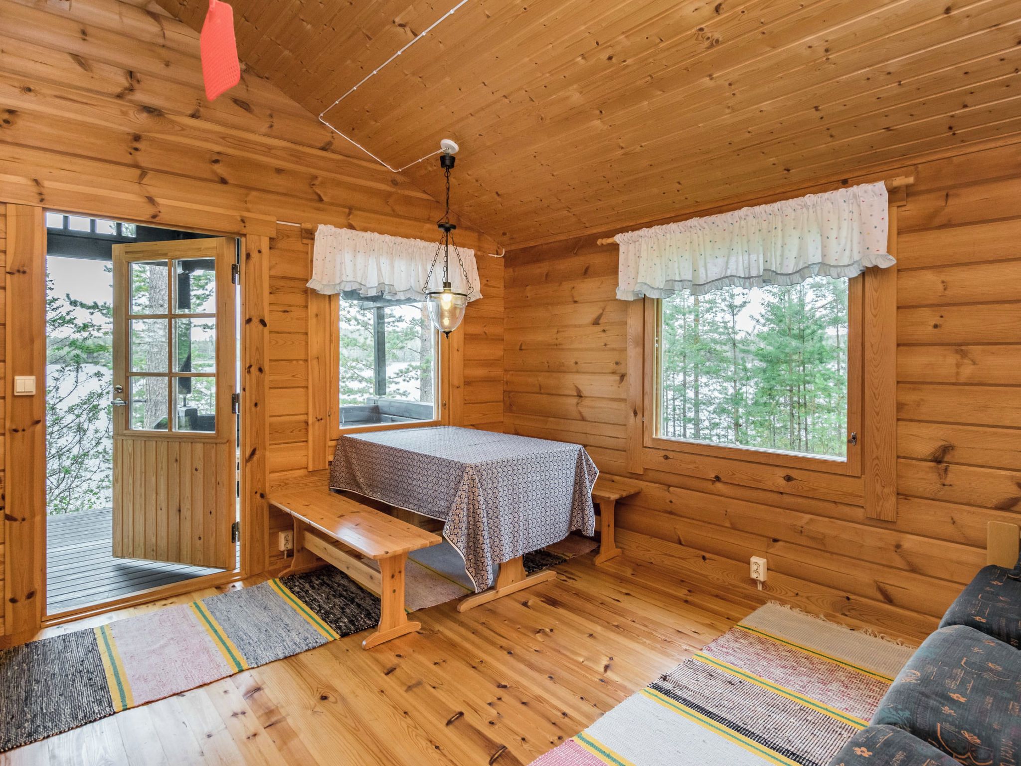 A wooden cabin interior with a bench, table, and two windows with curtains.