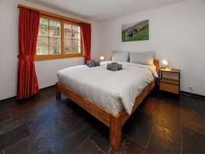 Een slaapkamer met een houten bed, rode gordijnen, een nachtkastje met lamp en een schilderij van een koe aan de muur.