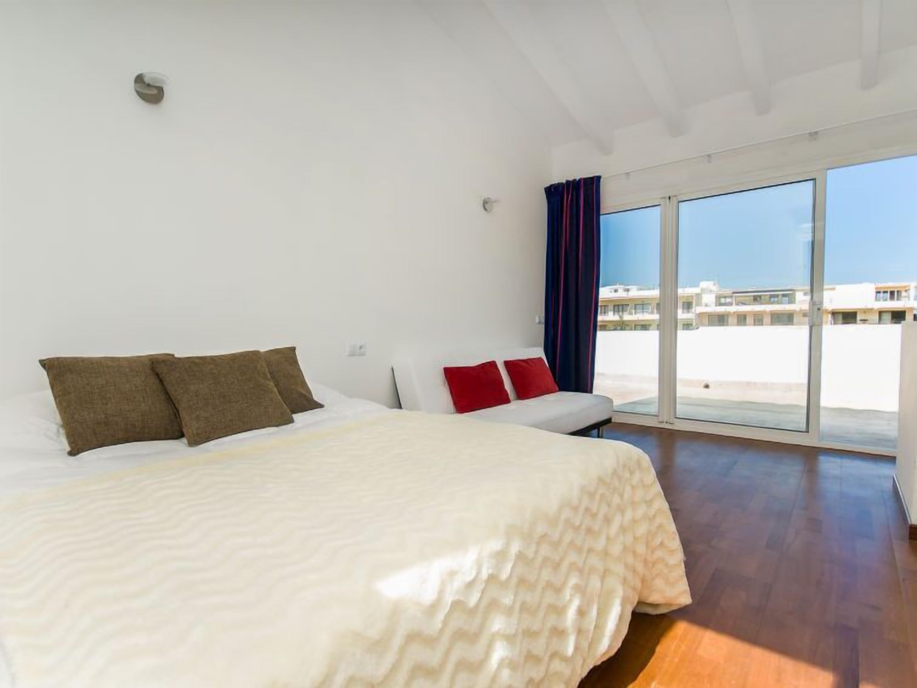 A modern hotel room with a large bed, a sofa, and a view of the city through large windows.