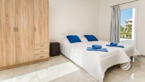 Ein Schlafzimmer mit großem Holzschrank, einem Bett mit weißer Bettwäsche und blauen Kissen sowie Blick auf ein Gebäude durch ein Fenster.