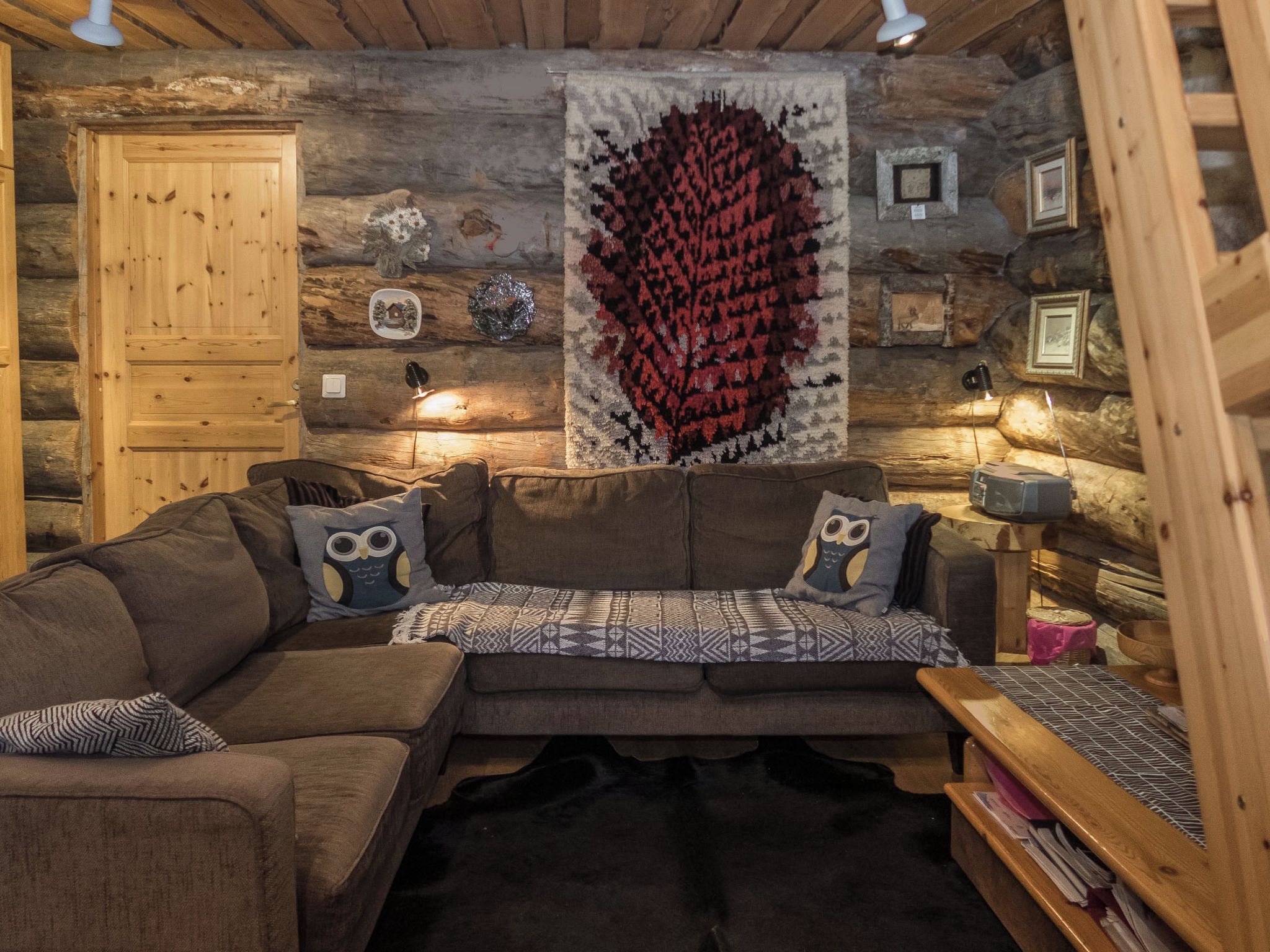 A cozy log cabin interior with a sectional sofa, two owl-themed pillows, a patterned throw, and a large red and black tapestry.