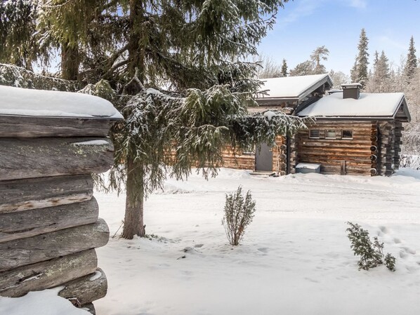 A log cabin with a snow-covered roof, surrounded by snow-laden trees and a clear sky.