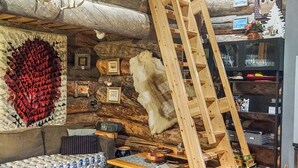 Tunnelmallinen, maalaishenkinen mökin sisustus, jossa on puinen tikapuu, sohva, takka ja seinä, joka on koristeltu kehystetyillä kuvilla ja suurella punaisen lehden taideteoksella.
