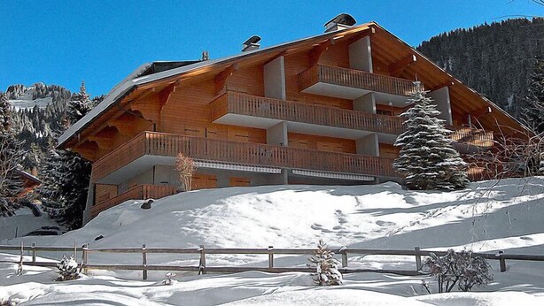 Een houten chalet met meerdere verdiepingen en balkons, met een besneeuwde berg op de achtergrond.