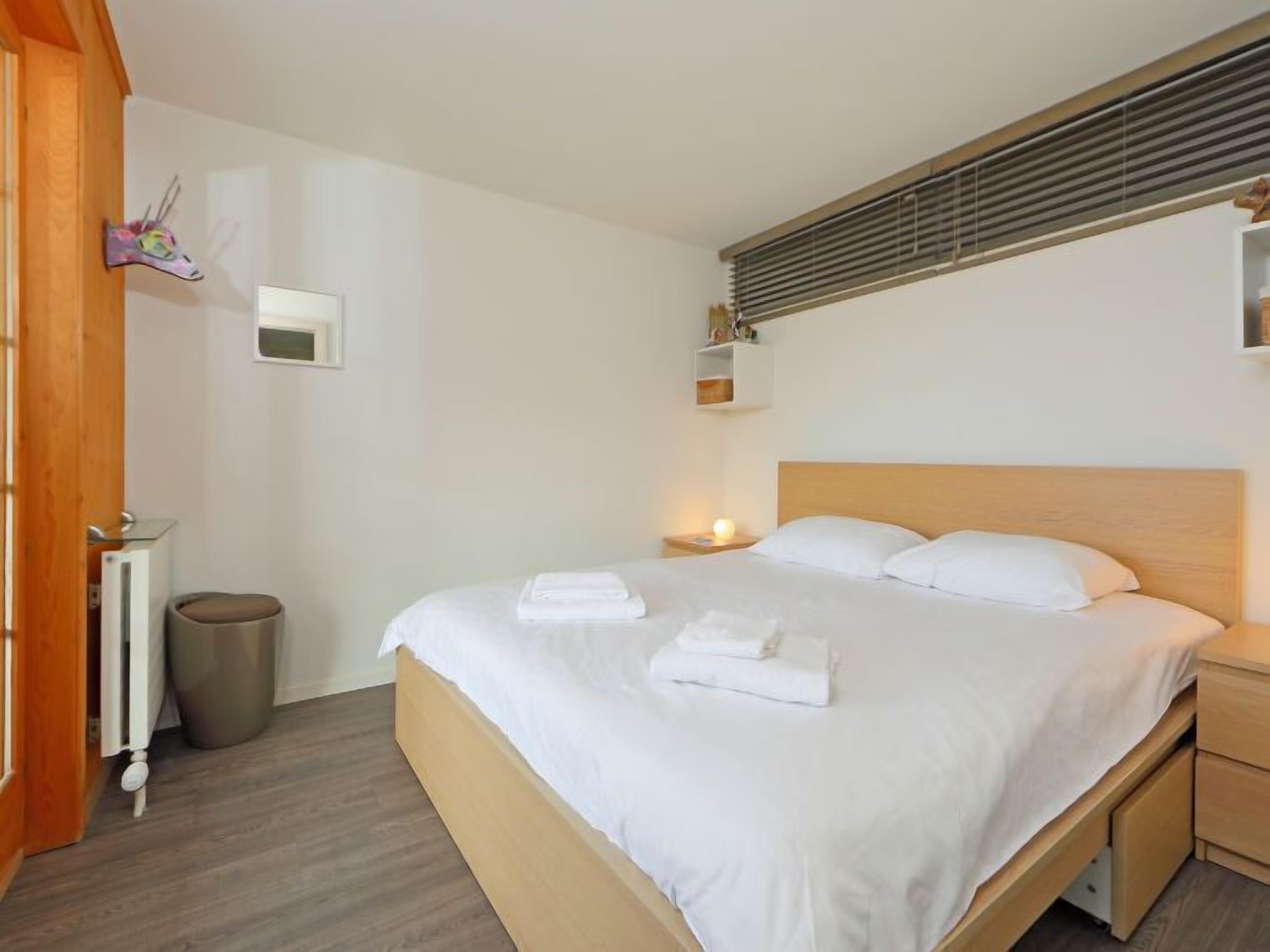 A hotel room with a large bed, wooden headboard, a bedside table with a lamp, a small trash bin, and a radiator.