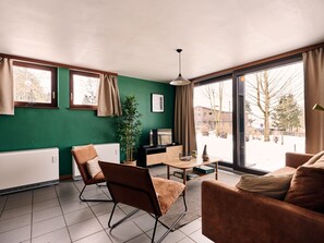Ein modernes Wohnzimmer mit grüner Wand, Heizkörper, Sofa, Sessel, Tisch, Pflanze und Fernseher.
