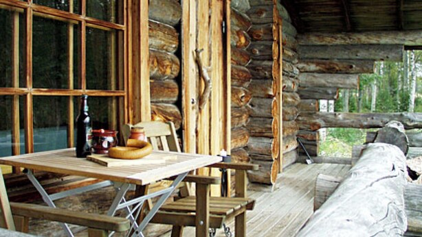 A rustic wooden cabin with a wooden table and chairs, a window with a view of trees, and a log wall.