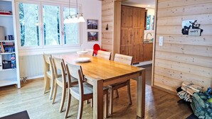 Ein Zimmer mit Holzwänden, einem Esstisch, Stühlen, einem Heizkörper und einem Fenster mit Spitzenvorhängen.