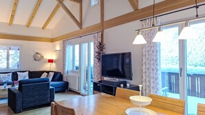 Ein geräumiges Wohnzimmer mit Holzbalkendecke, einem Fernseher, einem Esstisch mit Stühlen und großen, mit Vorhängen versehenen Fenstern.