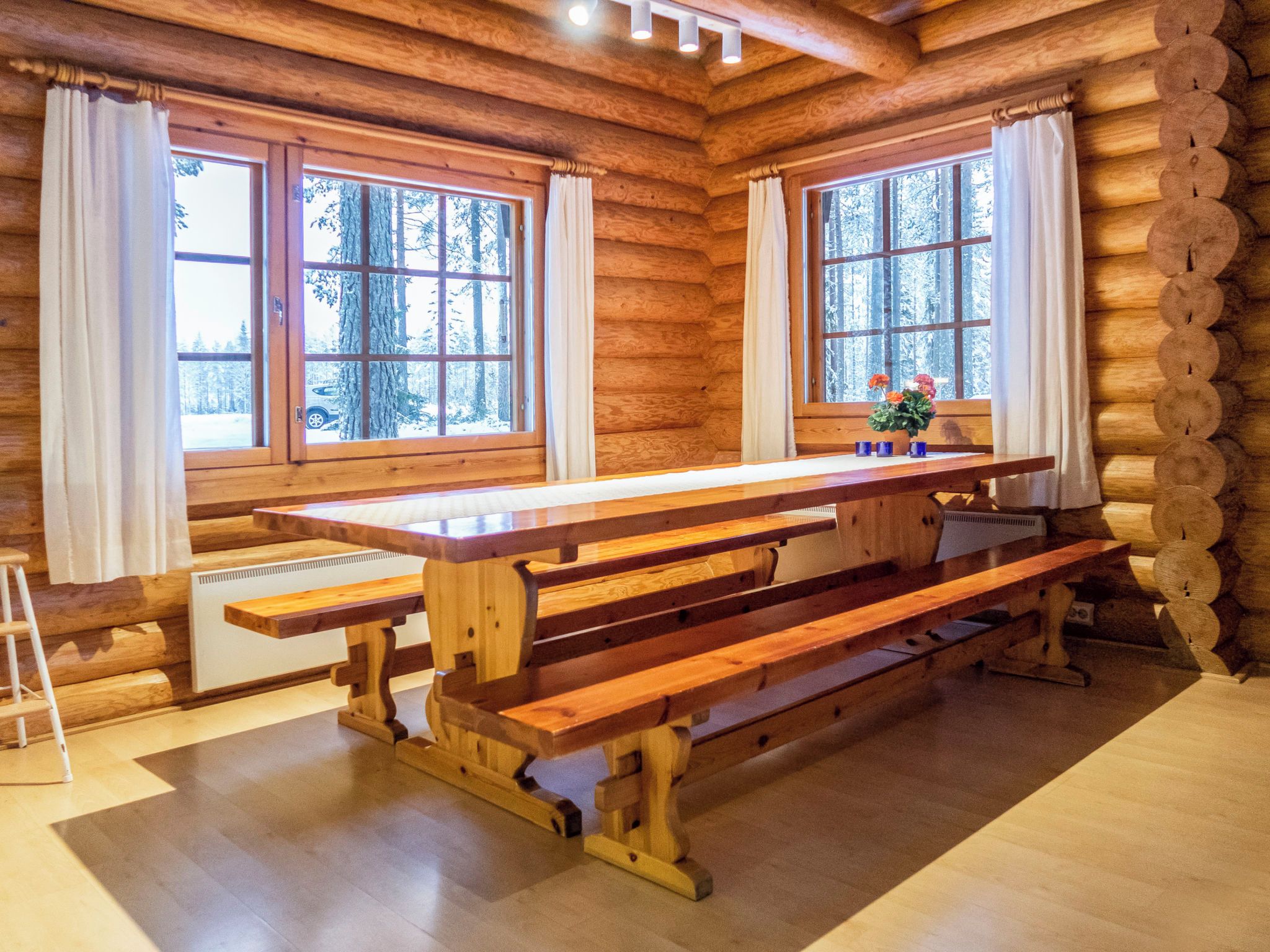 A wooden cabin interior with a large dining table, two windows with curtains, and a view of a snowy forest.