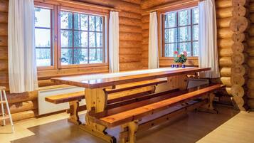 A wooden cabin interior with a large dining table, two windows with curtains, and a view of a snowy forest.