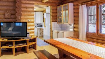 Ein Holzblockhaus-Interieur mit Fernseher, Holzmöbeln, einer Küchenzeile und Blick auf die verschneite Landschaft.