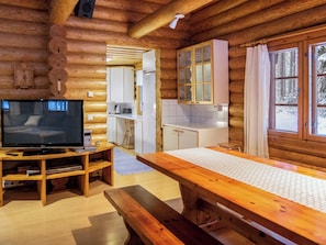 A log cabin interior with a television, wooden furniture, a kitchen area, and a view of the snowy outdoors.