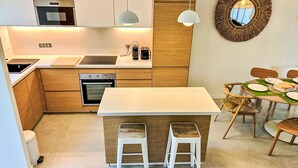 A modern kitchen with wooden cabinets, a central island with stools, a dining area with a round table, and a decorative mirror on the wall. - Les Restanques Golfe Saint Tropez by Interhome (Grimaud)