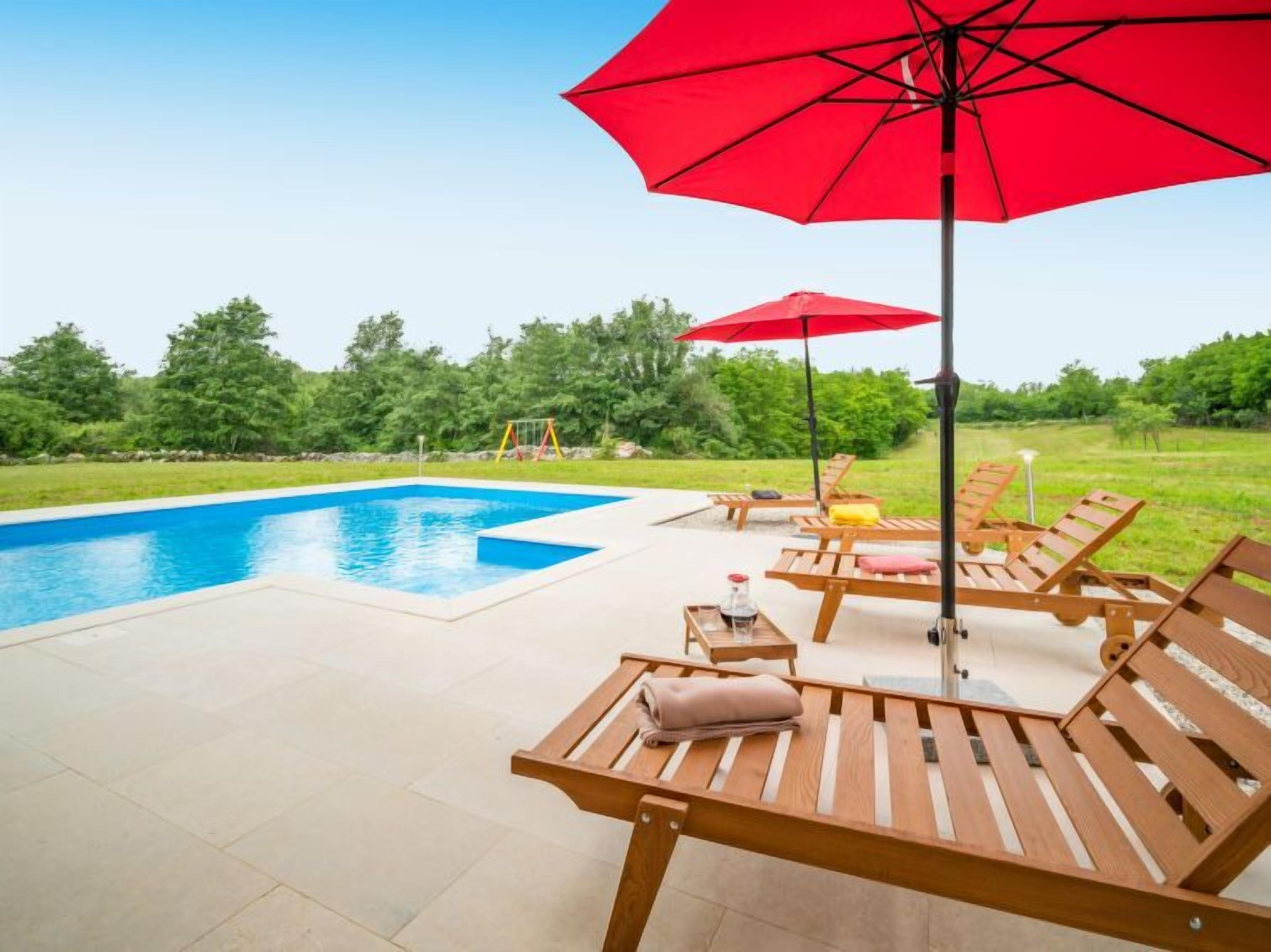A swimming pool with wooden lounge chairs and red umbrellas.