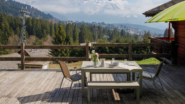 A wooden deck with a table and chairs, overlooking a mountainous landscape with a view of snow-capped peaks and a town below. - Saint Piran by Interhome (Villars)
