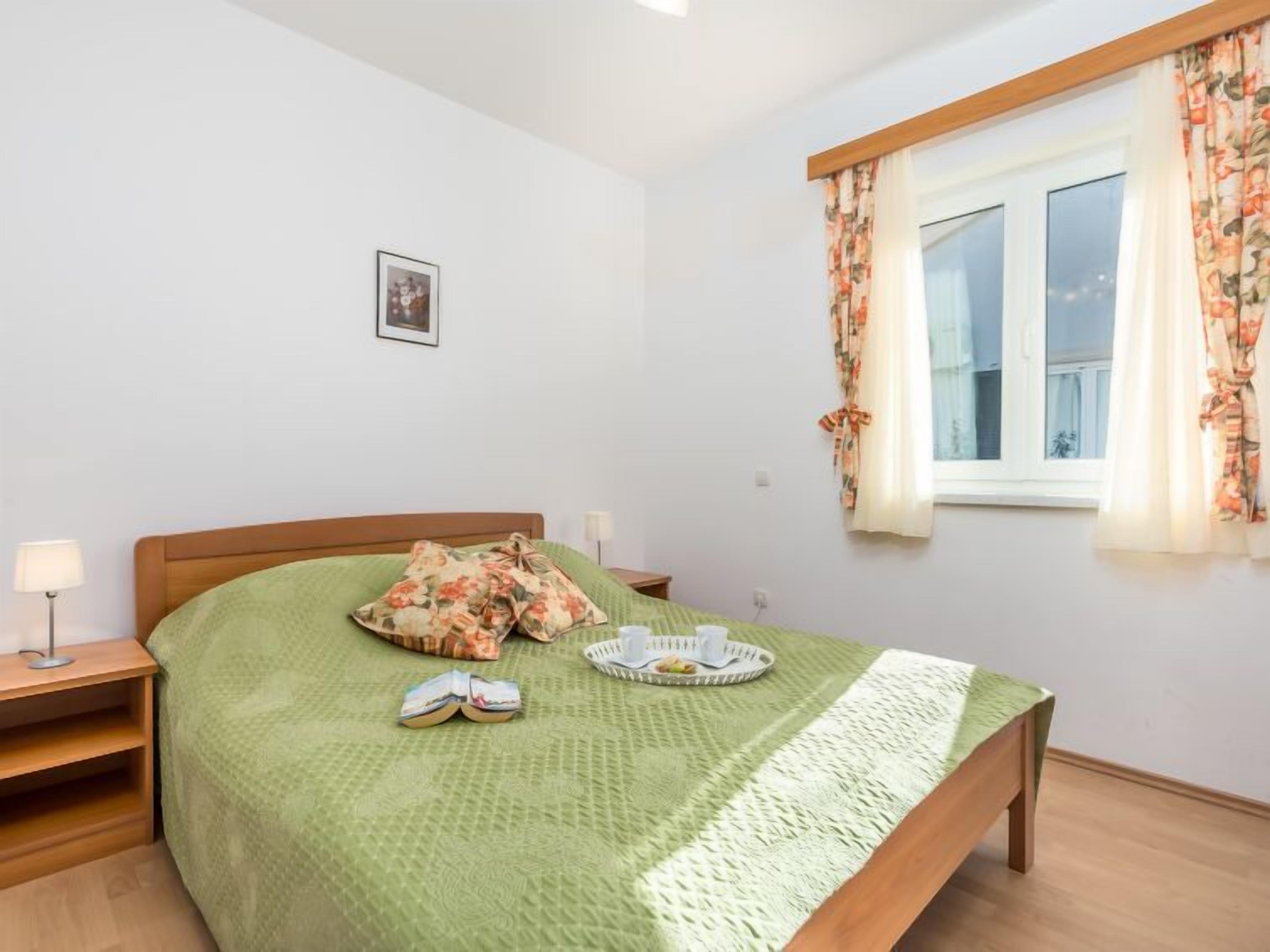 A neatly arranged bedroom with a bed, a bedside table, a lamp, a window with curtains, and a small picture on the wall.