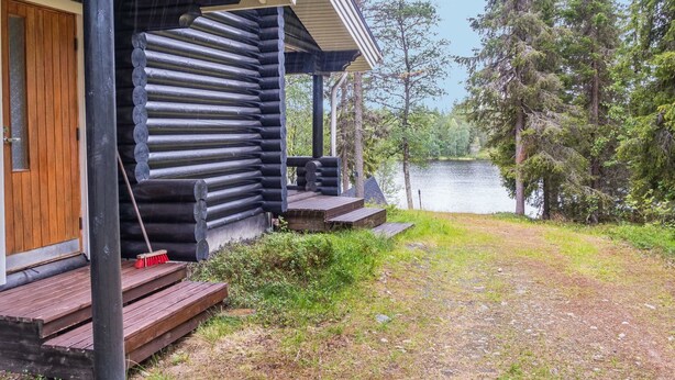 A log cabin with a porch, steps leading to a wooden door, and a view of a lake surrounded by trees.