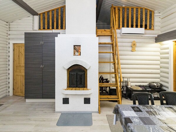 A log cabin interior with a central fireplace, wooden staircase, and a dining area with tables and chairs.